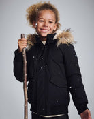 A child wearing a Black City Transit Coat with faux fur hood, smiling while holding a stick in the snow.