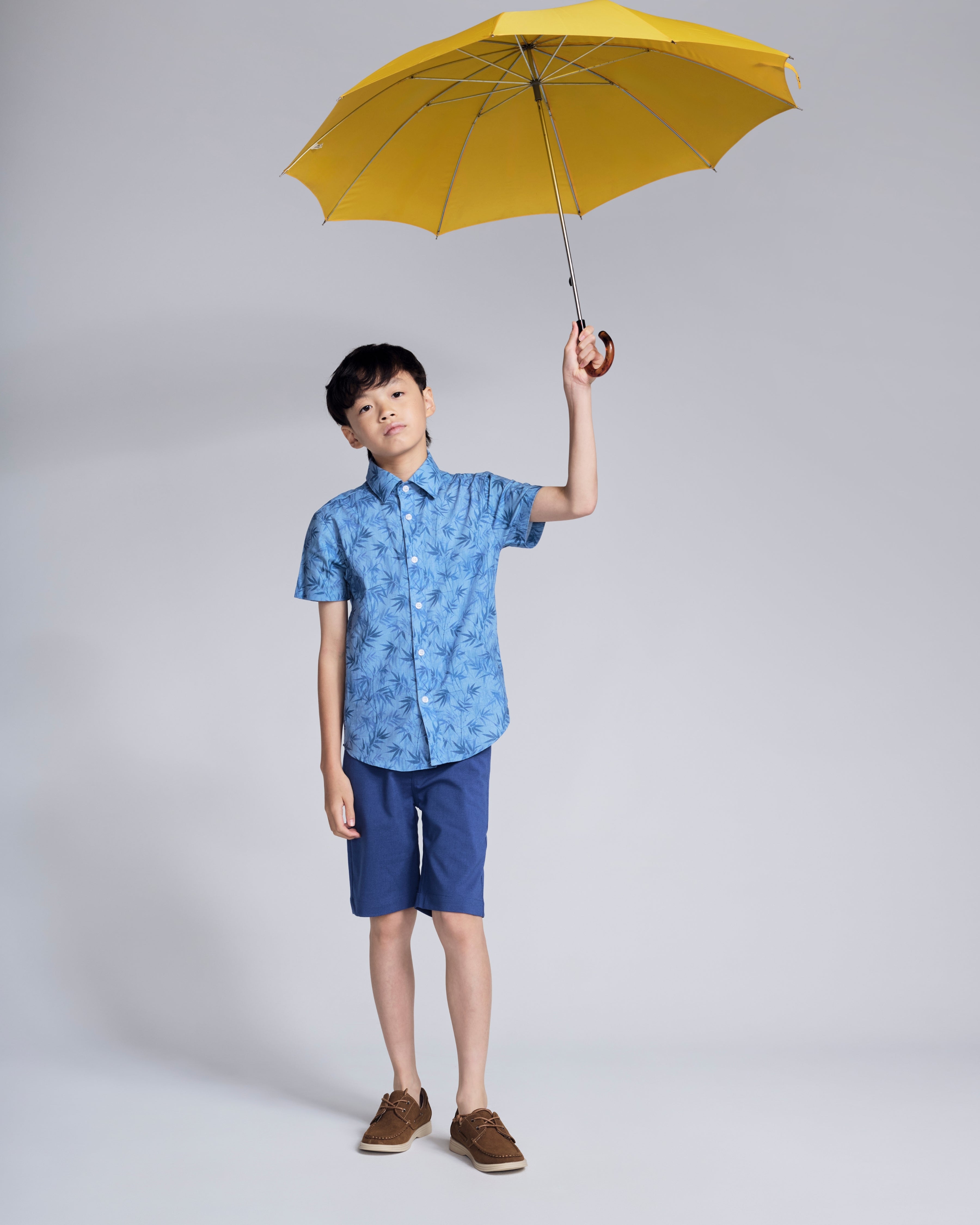 A child wearing a Blue Bamboo Day Party Shirt, holding a yellow umbrella, showcasing casual summer style.