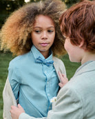 Two children standing close to each other outdoors, one wearing a light blue shirt with a bow tie.
