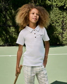Child wearing a white polo shirt with brown buttons on a tennis court