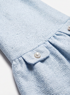 Close-up of a light blue textured fabric garment detail, showing a pleated section, a small flap pocket with a pearl button, and part of a matching pearl button on the sleeve.