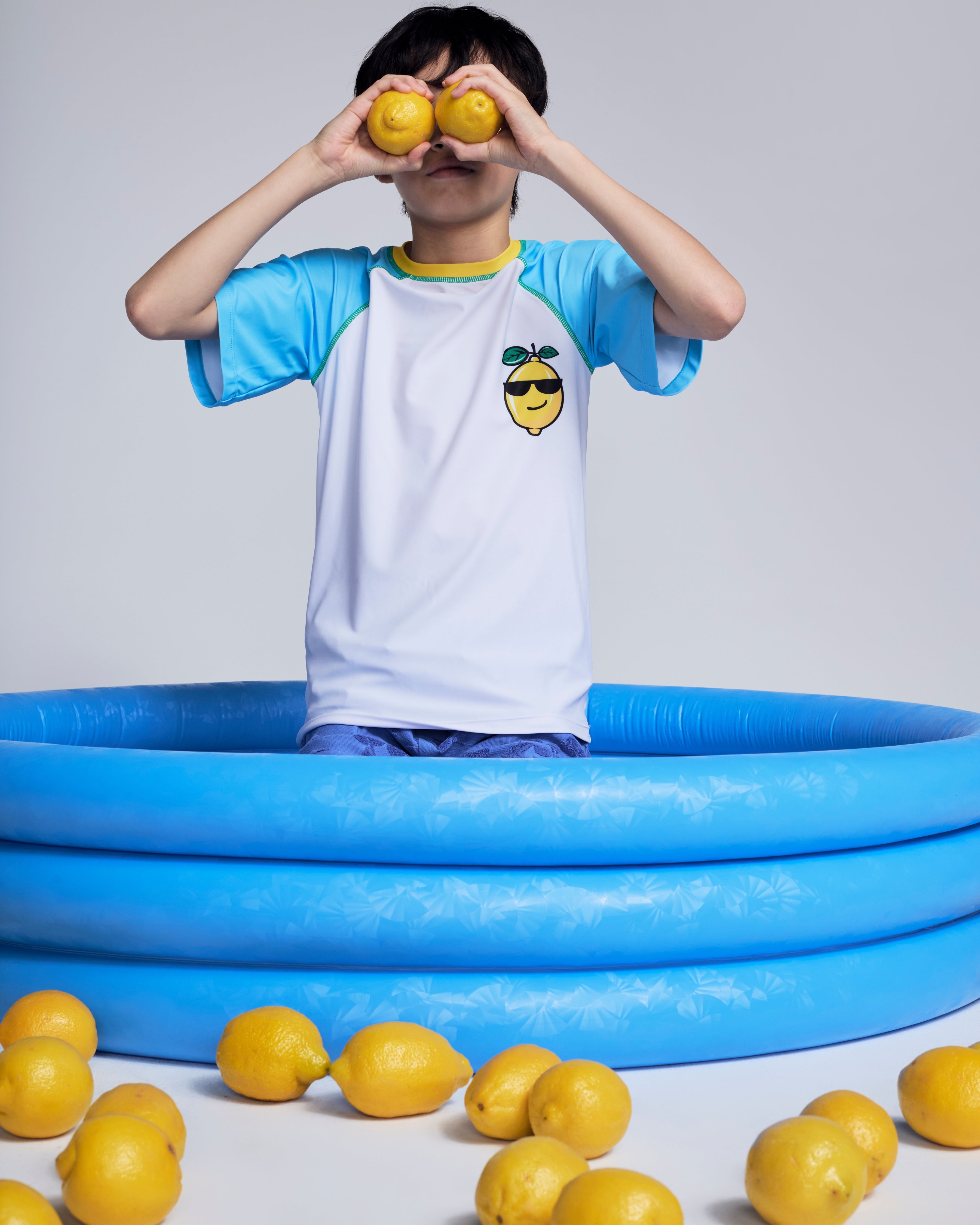 Child wearing a Cool Lemon Rash Guard, holding lemons, standing in a blue pool surrounded by lemons.
