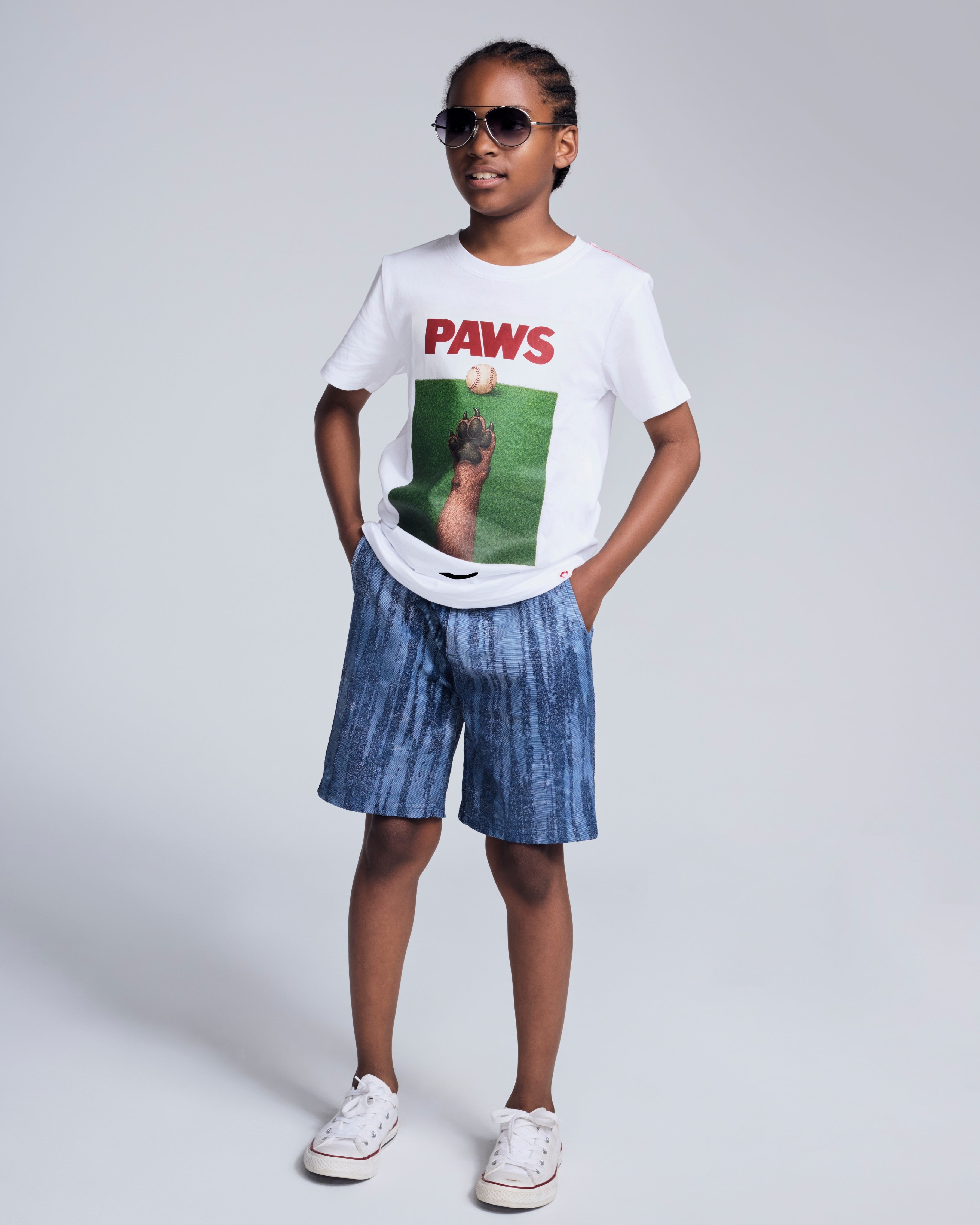 Kid wearing Dog Paws White Graphic Short Sleeve Tee with sunglasses and denim shorts on a grey backdrop.