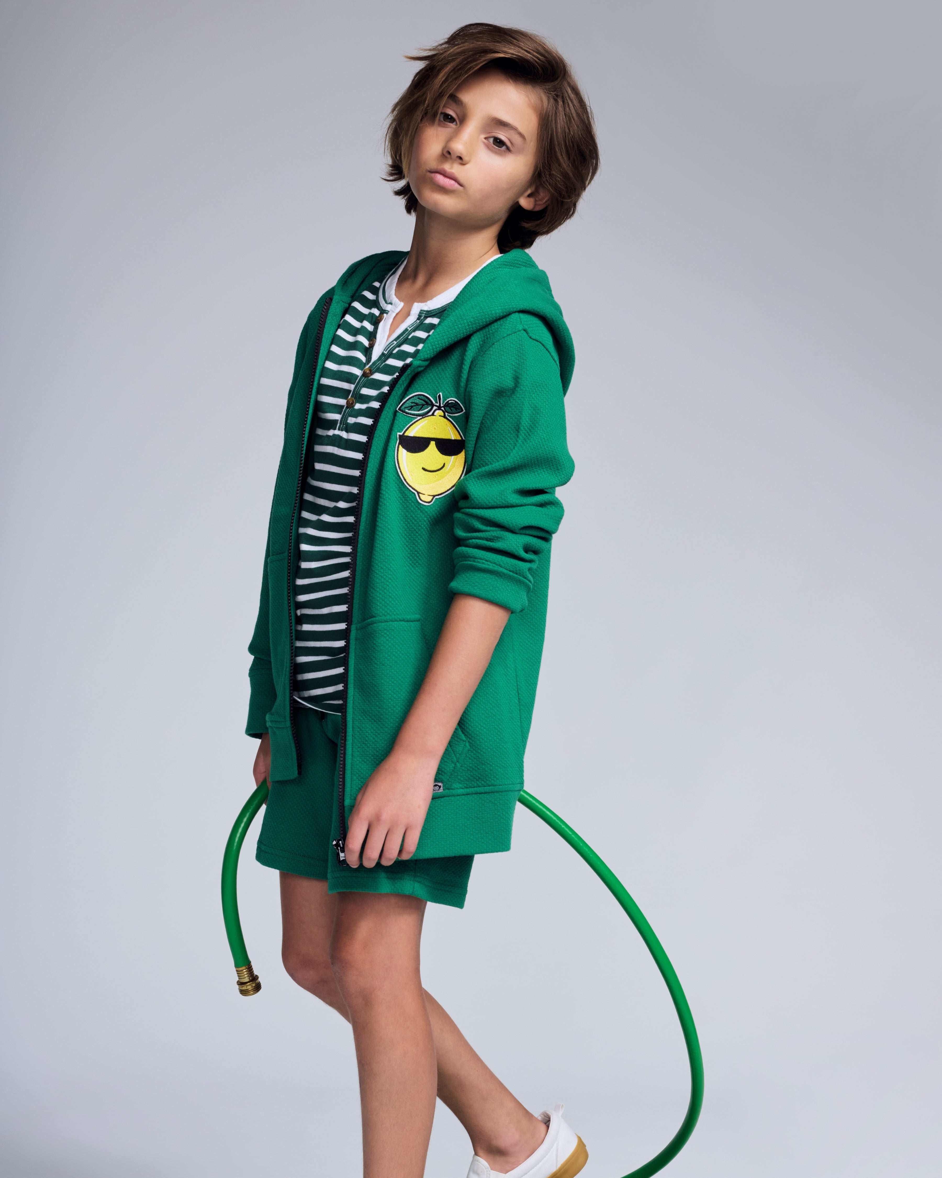 Boy wearing a Garden Green Resort Short with a green hoodie and striped shirt, holding a garden hose.