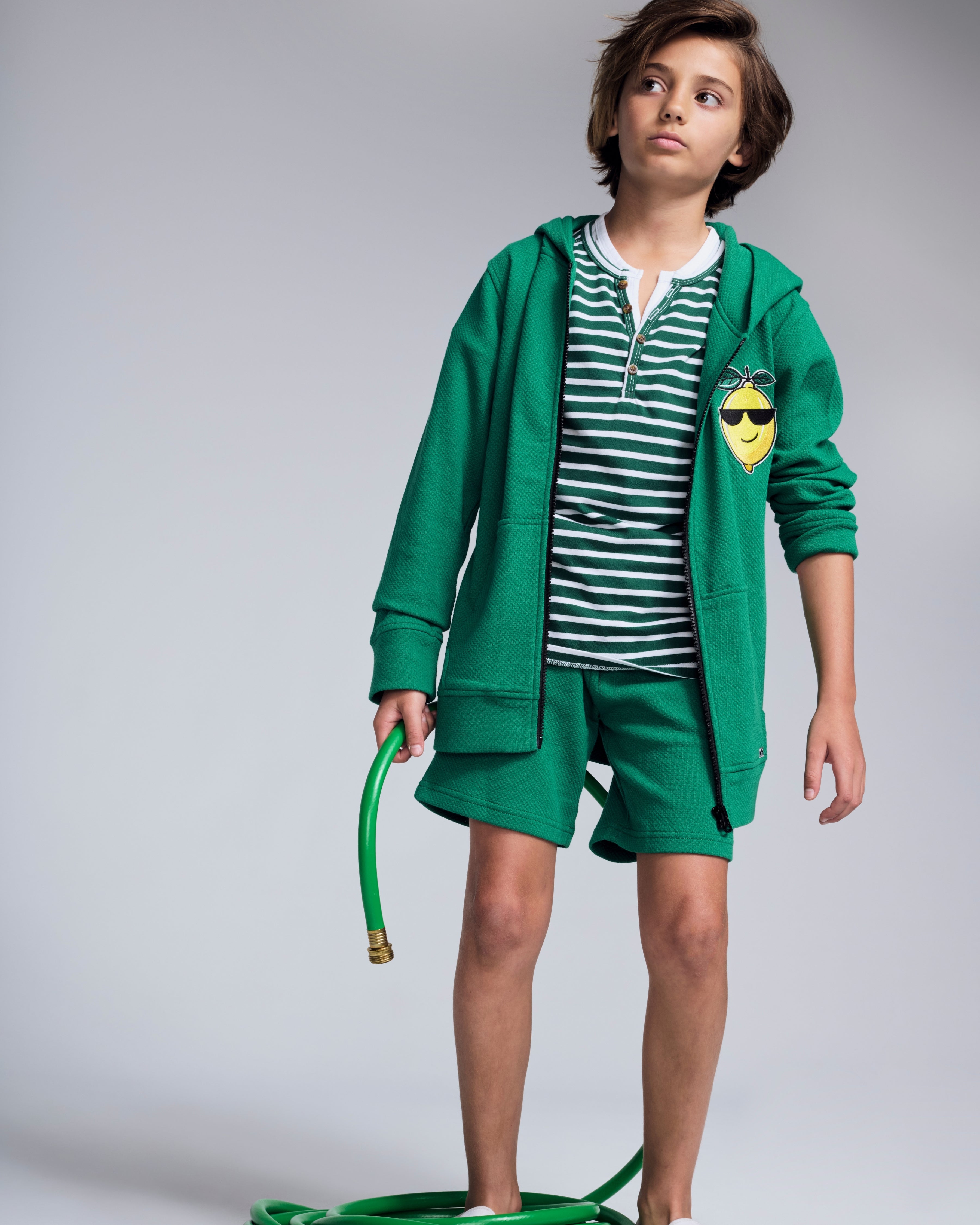 A child wearing the Garden Green Stripe Hilltop Henley with green shorts and a hoodie, holding a garden hose.