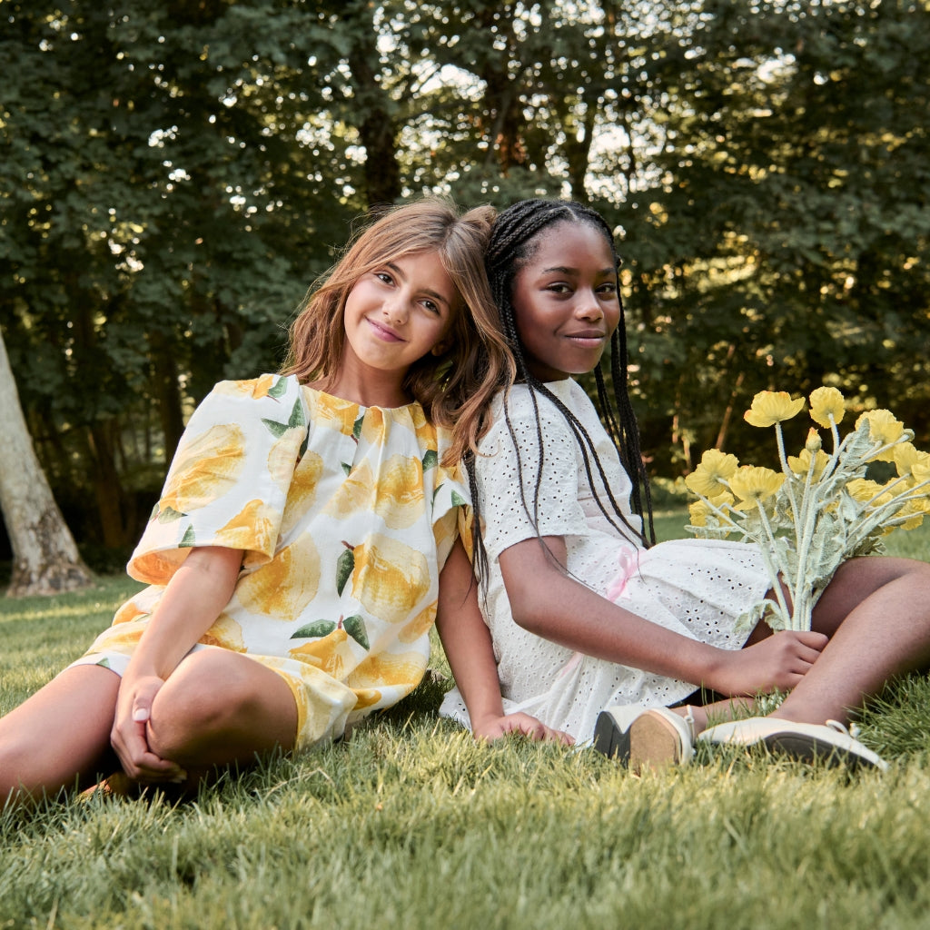 Two young girls sitting on grass holding yellow flowers, surrounded by greenery.