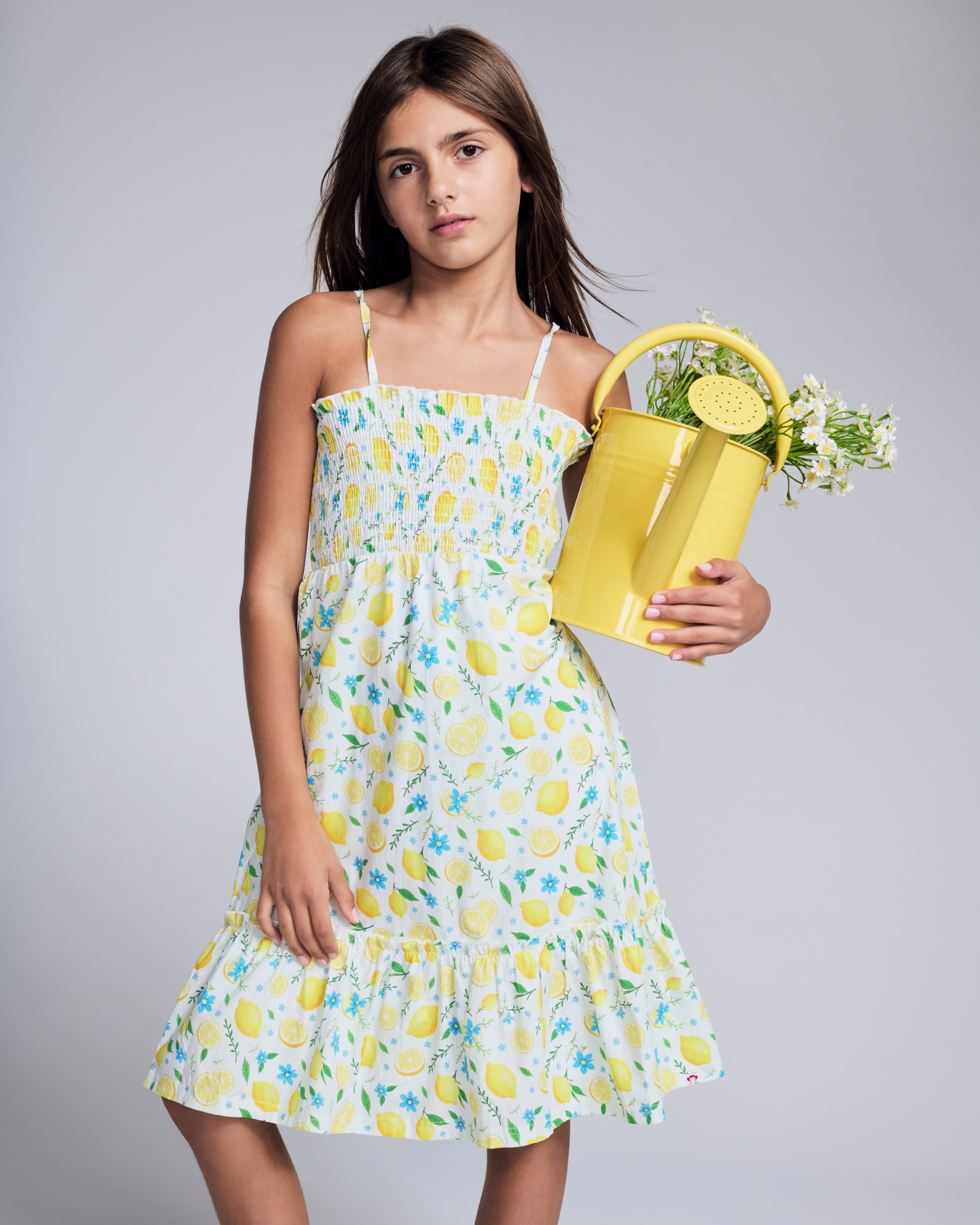 Girl wearing a Lemon Zest Madison Dress holding a yellow bag with flowers, showcasing ruffles and a lemon print.