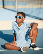 Chill kid rocking denim blue Resort Shirt with palm trees, sunglasses, and a cool vibe on the tennis court.