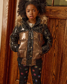 Child wearing a shiny puffer jacket with fur hood in front of a wooden door.