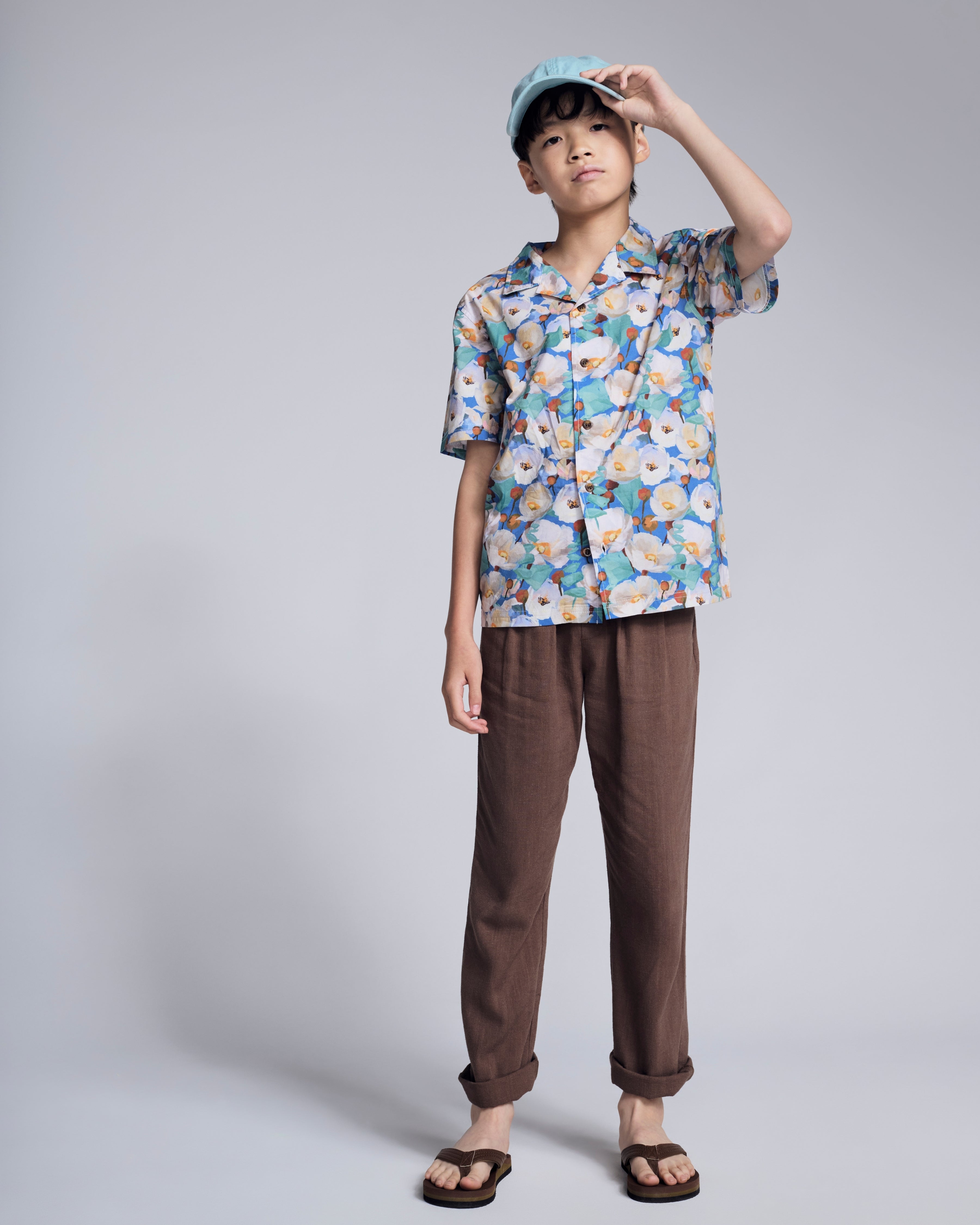 A young boy wearing a colorful floral shirt, brown pants, and a light blue cap, showcasing the Linen Brown Resort Pant.