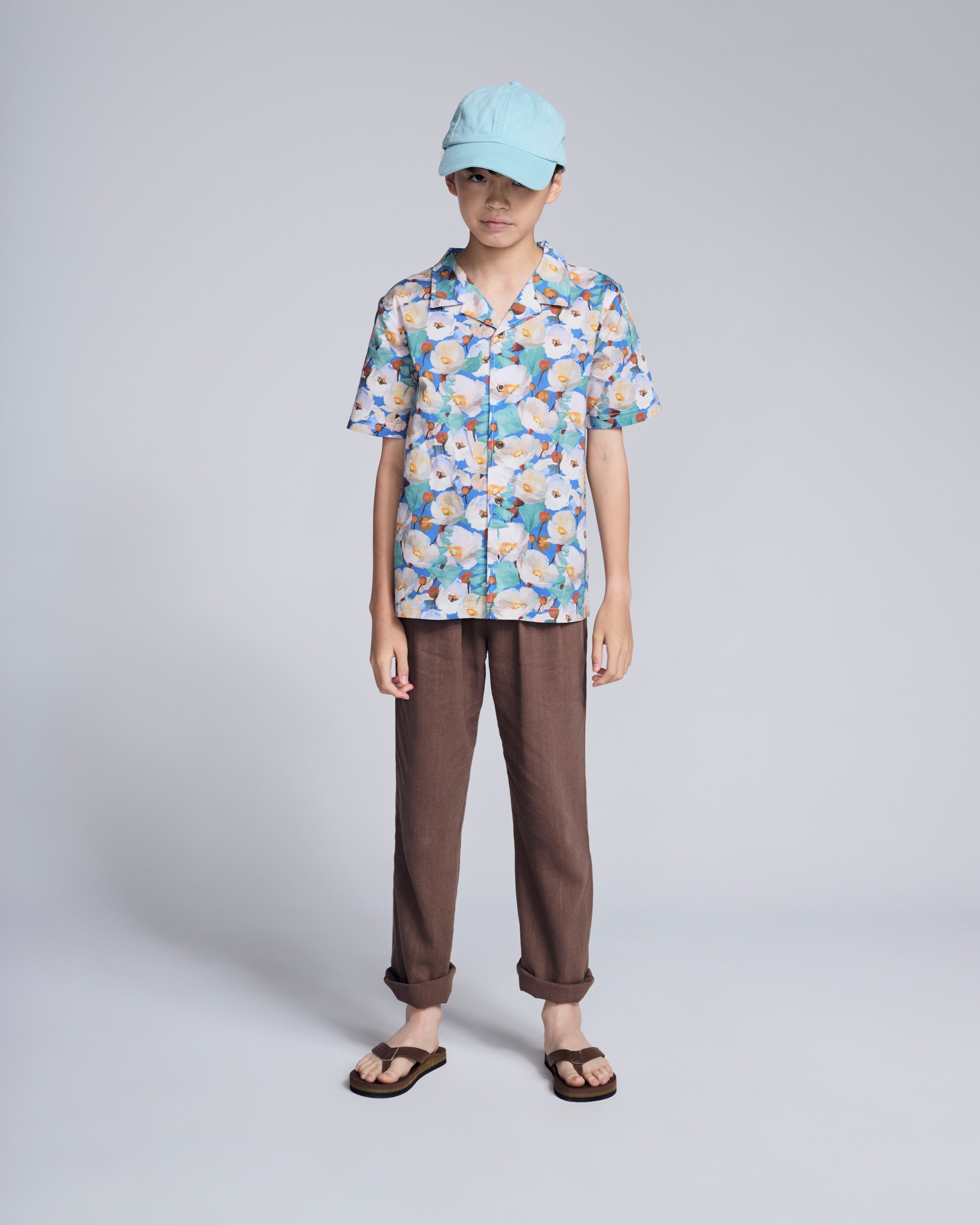 Child wearing a floral shirt and Linen Brown Resort Pant, styled with a blue cap and sandals.