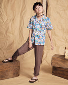 Boy modeling the Linen Brown Resort Pant with a colorful floral shirt and sandals on a wooden backdrop.