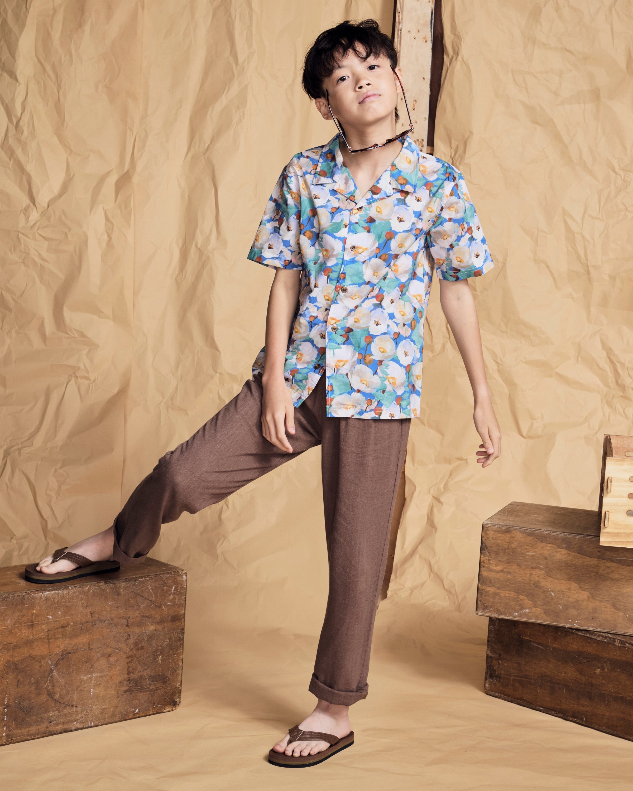 Boy modeling the Linen Brown Resort Pant with a colorful floral shirt and sandals on a wooden backdrop.