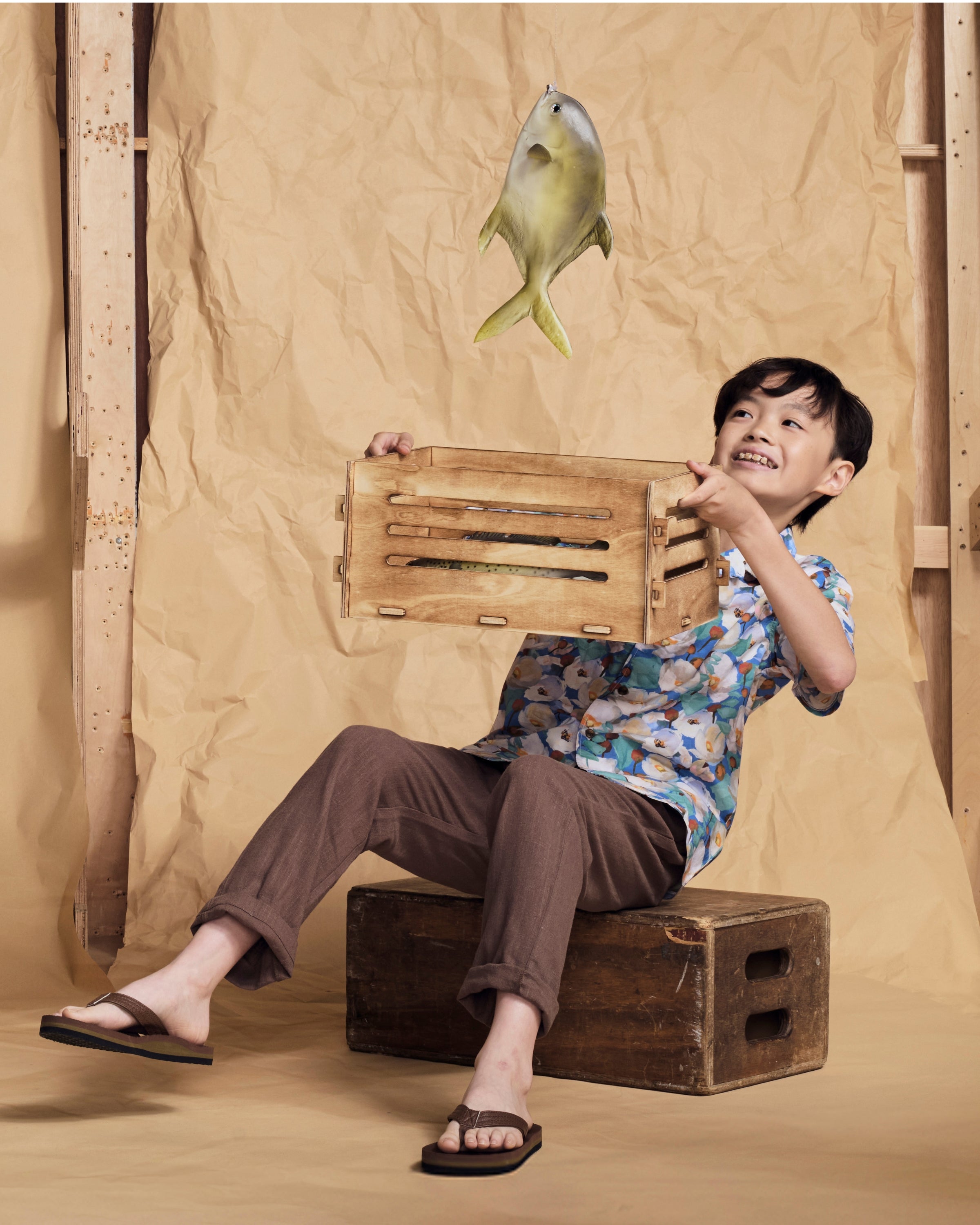 Boy in floral shirt and Linen Brown Resort Pant catches fish with wooden crate, playful summer scene.