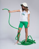 Girl in Polaroid White Circle Tee and green bike shorts holding a garden hose.