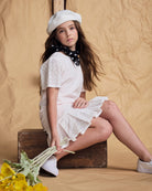 A girl wearing the Soft Pink Eyelet Eve Dress with a pleated skirt, sitting on a wooden box holding flowers.