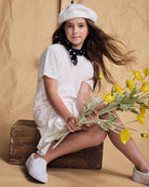 Girl wearing Soft Pink Eyelet Eve Dress with flowers, styled with a beret and scarf, set against a warm background.