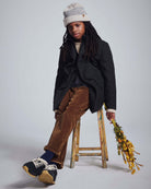Child in stylish coat and hat wearing British Khaki Cord Club Pants, sitting on a stool with flowers.