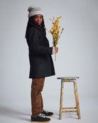 Young model wearing British Khaki Cord Club Pants, gray coat, holding flowers, styled with a playful hat.