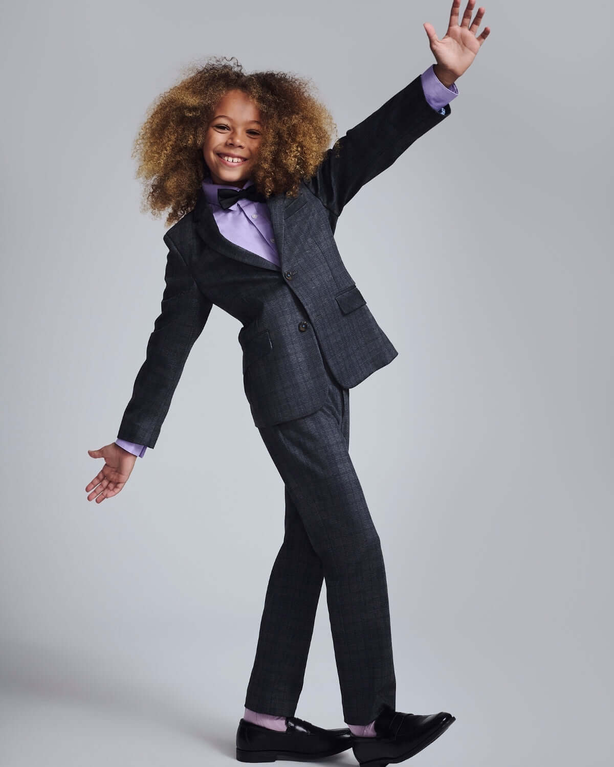 A young boy in Gentleman's Plaid Stretchy Suit Pants and a bow tie, smiling and posing in a stylish manner.