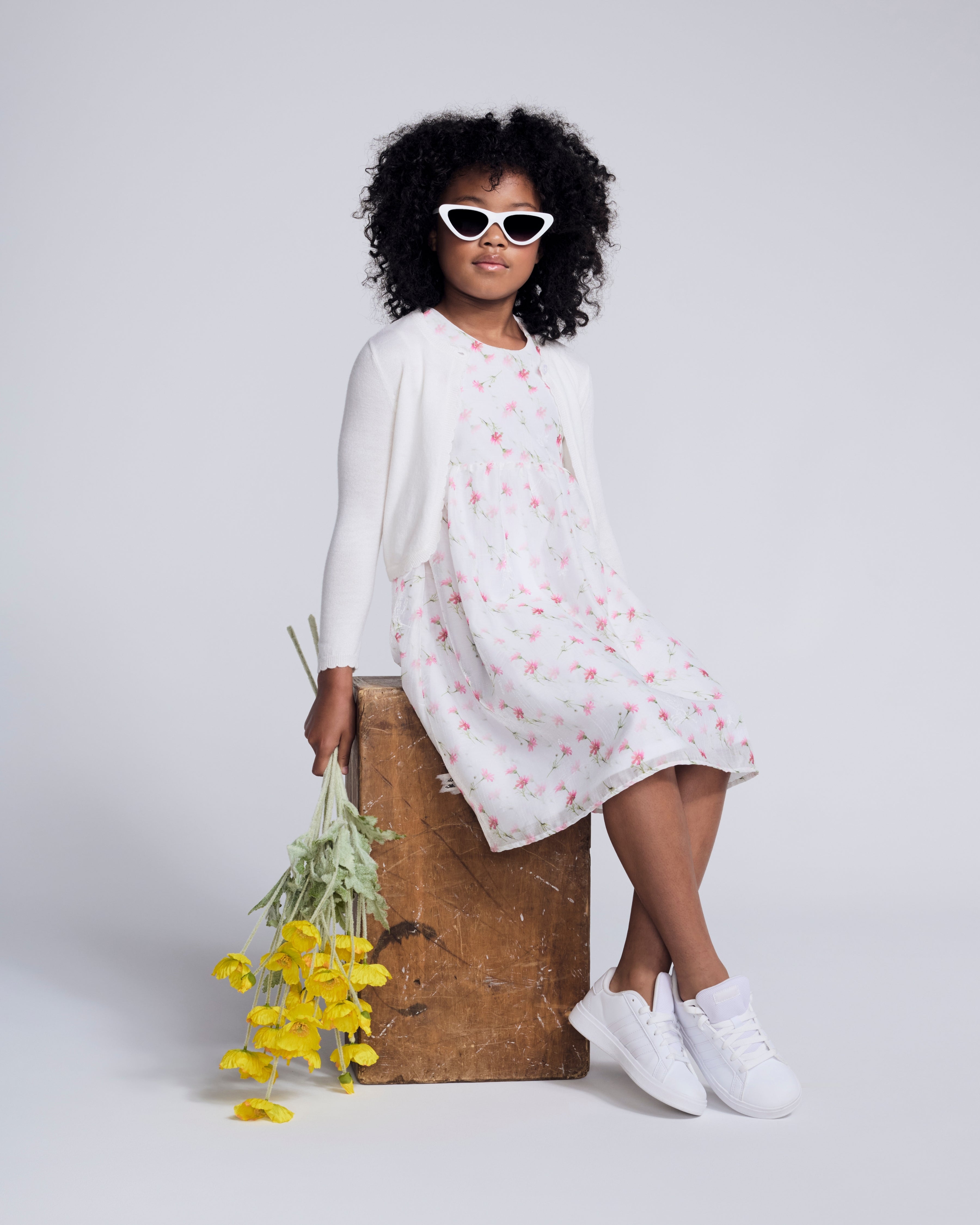 Girl wearing a White Bolero Cardigan over a floral dress, accessorized with sunglasses and holding yellow flowers.