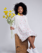 Girl wearing a White Rose Empire Dress, holding yellow flowers, sitting on a wooden crate.