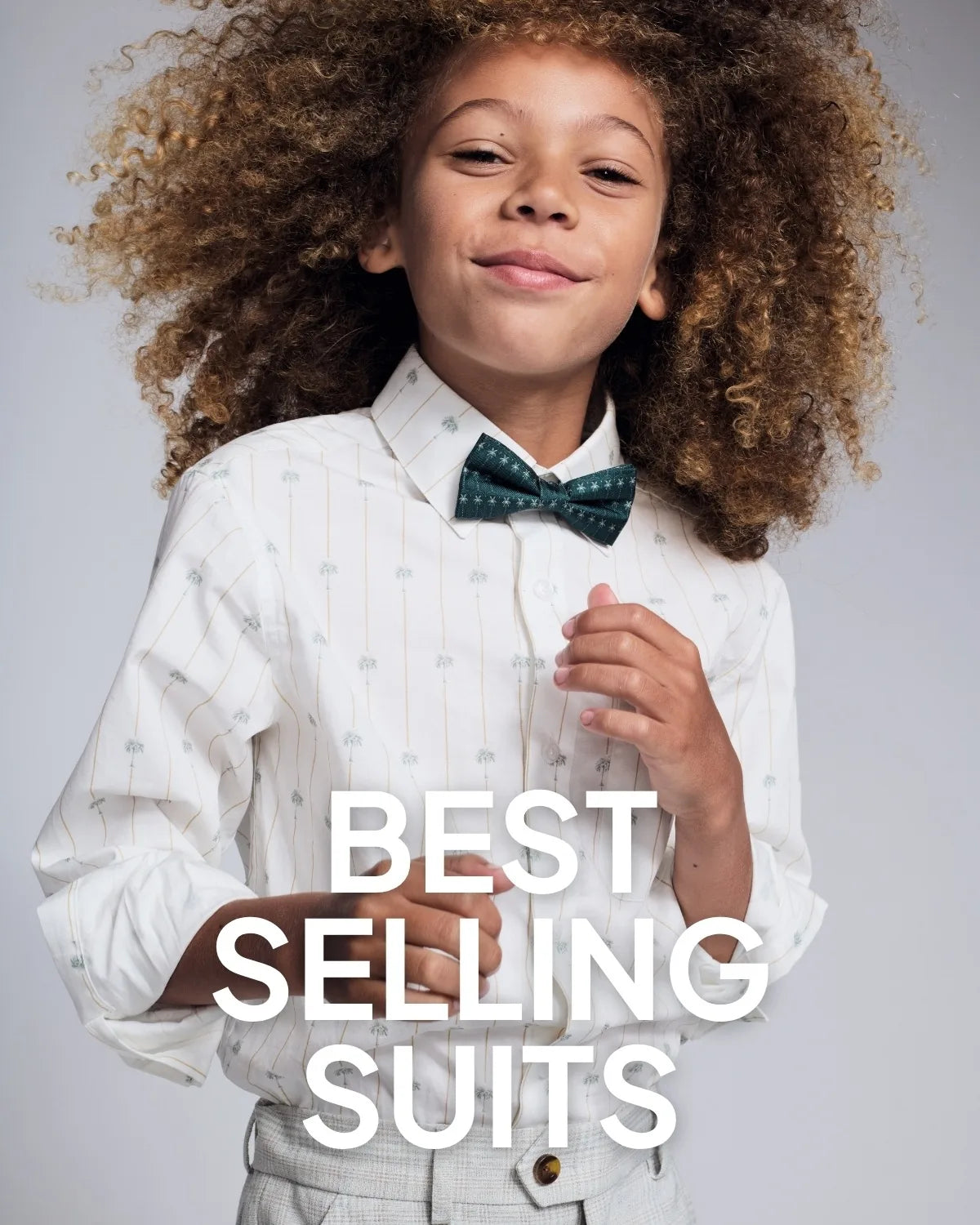 A young child with voluminous curly hair wearing a white pinstriped button-up shirt and a dark green patterned bow tie, posed against a light gray studio background with large white text reading 'BEST SELLING SUITS' across the lower center.