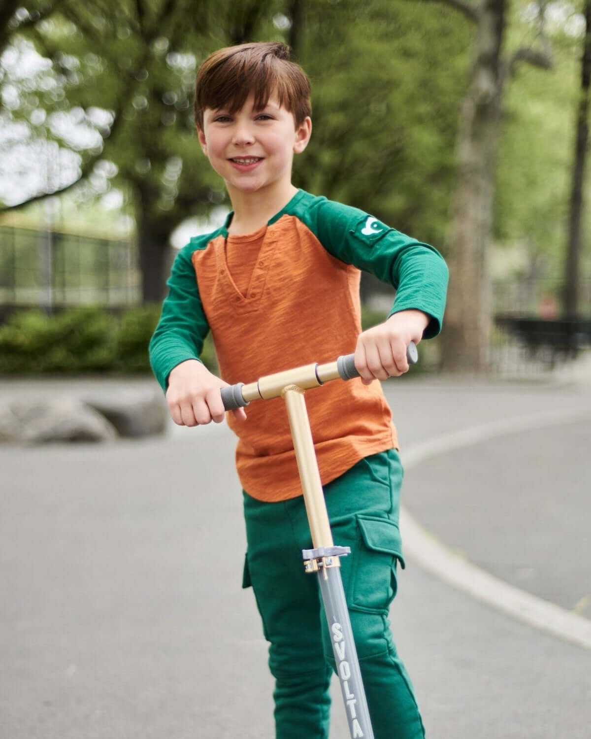 Boy riding a scooter wearing a Terracotta Baseball Tee and green sweatpants outdoors.