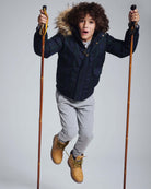 Child jumping in the Black Watch Tartan City Transit Coat with fur-lined hood and ski poles.
