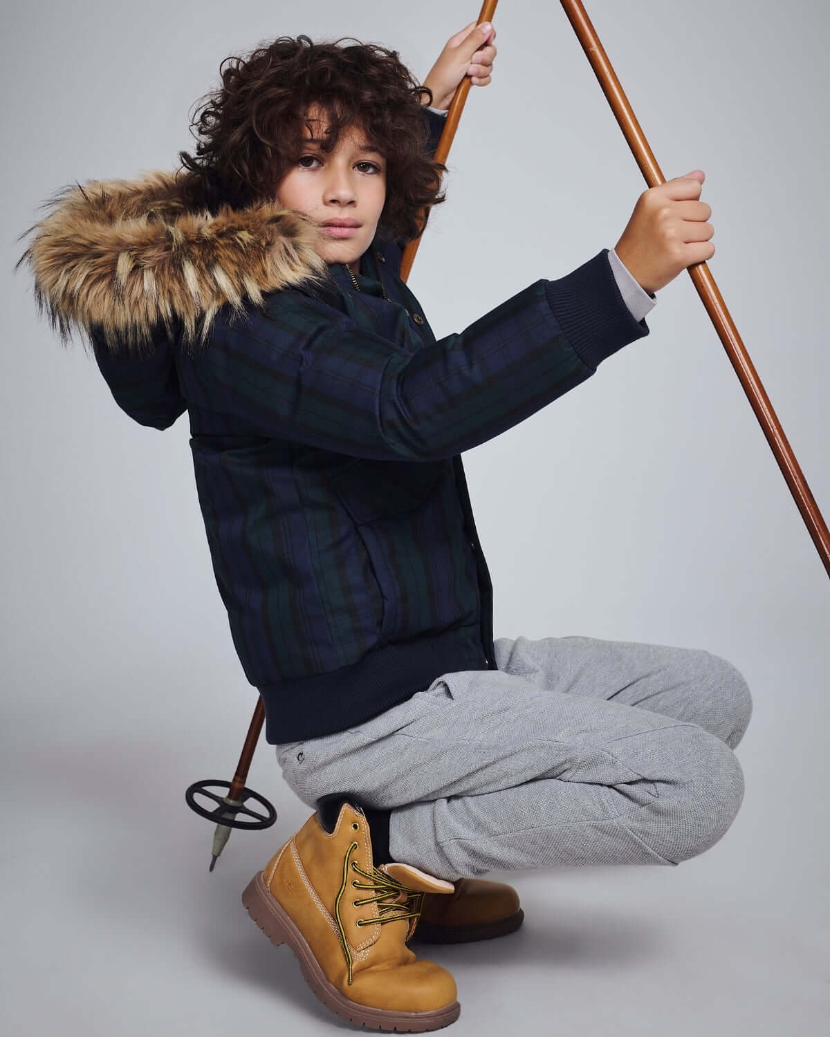 Child wearing Black Watch Tartan City Transit Coat with fur-lined hood and gray pants, posing with ski poles.