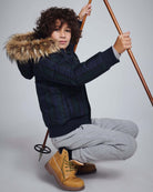 Child wearing Black Watch Tartan City Transit Coat with fur-lined hood and gray pants, posing with ski poles.