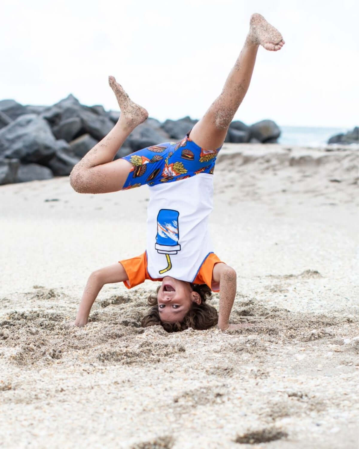 Child doing a handstand on the beach in Burgers & Fries Swim Trunks, showcasing fun summer style and playfulness.