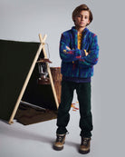 A young boy wearing Spruce Club Pants and a colorful jacket, standing in front of a tent for outdoor adventures.
