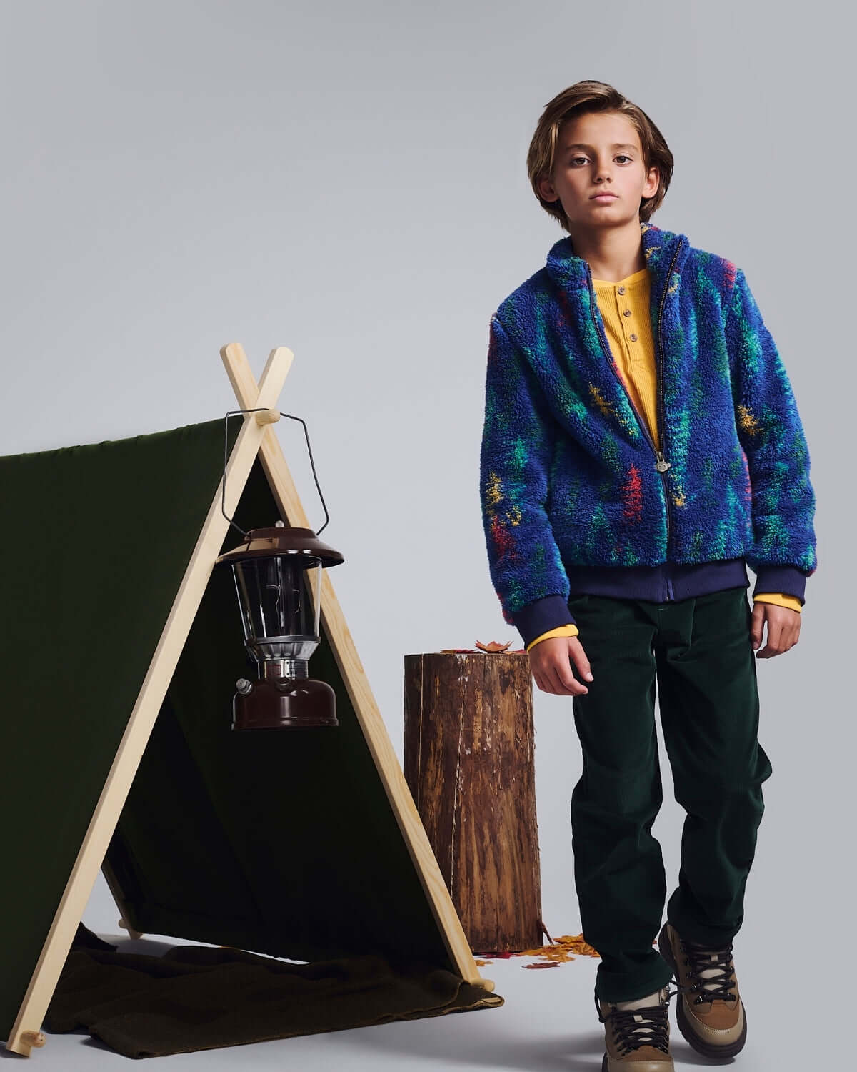 Boy wearing Spruce Club Pants and a colorful jacket next to a camping tent, showcasing adventure-ready style.