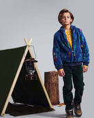 Boy wearing Spruce Club Pants and a colorful jacket next to a camping tent, showcasing adventure-ready style.