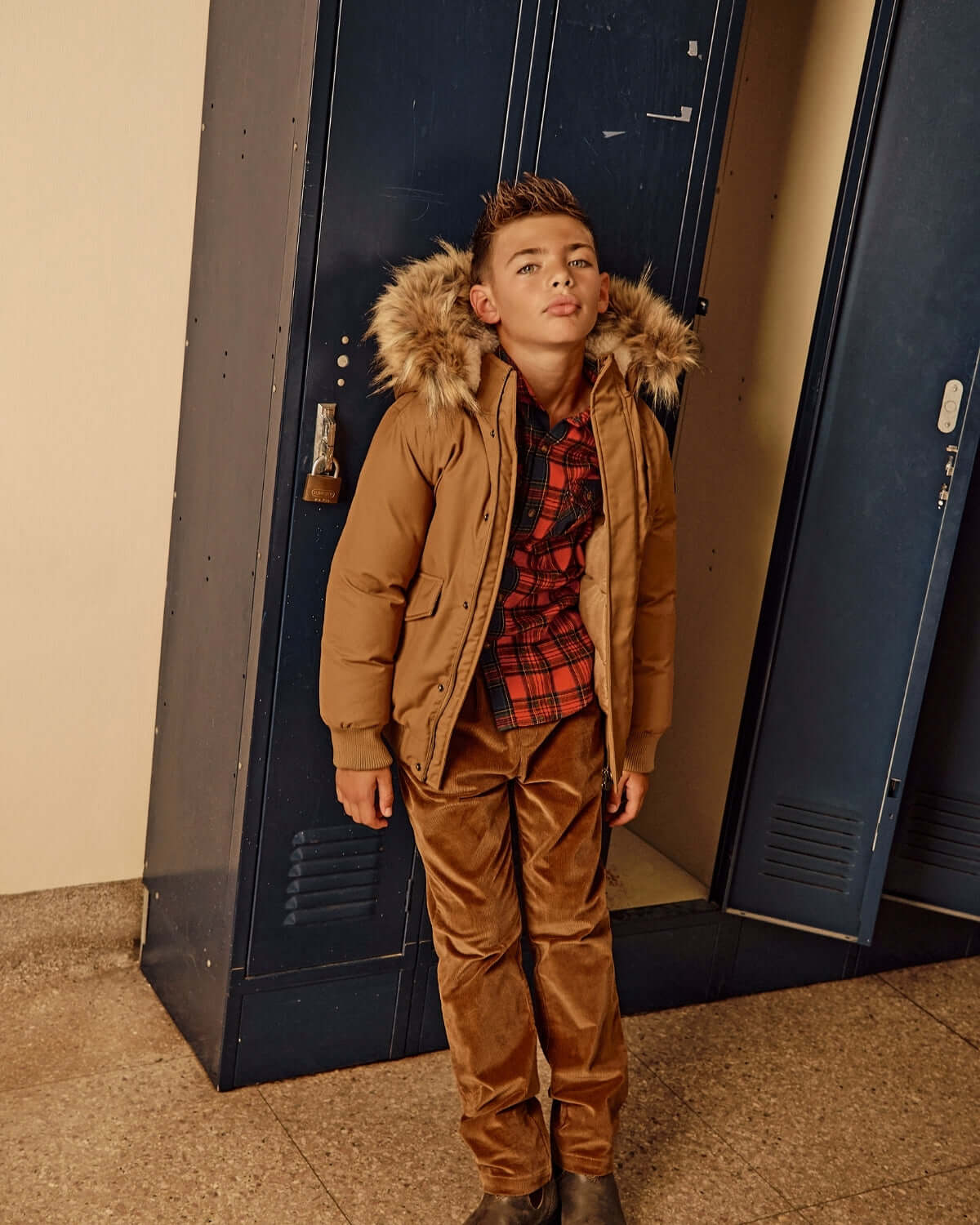 Boy wearing British Khaki Cord Club Pants with a brown jacket and plaid shirt, standing next to lockers.