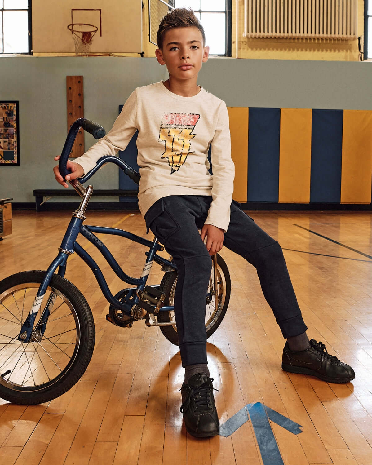 Boy wearing Creative Spark Graphic Long Sleeve Tee, sitting on bike in gym, showcasing bright lightning bolt design.
