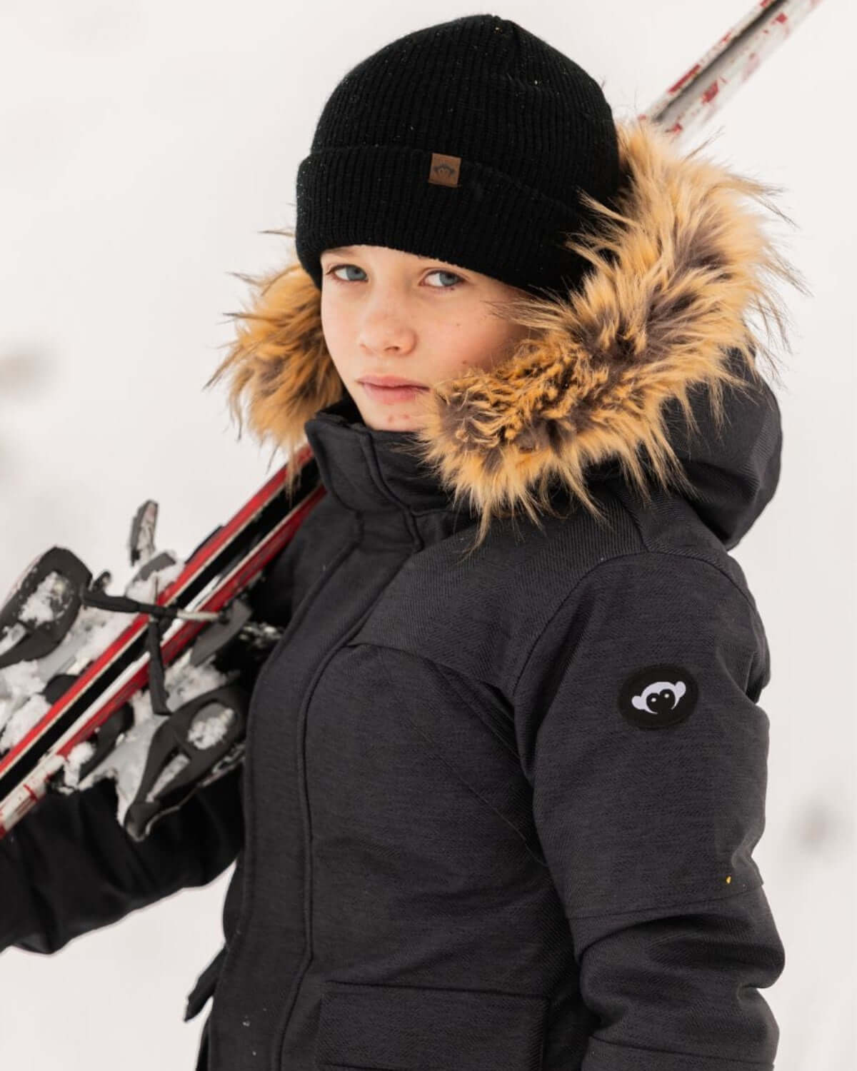 Stylish child wearing a Black Denali Down Coat while holding skis in a winter setting.