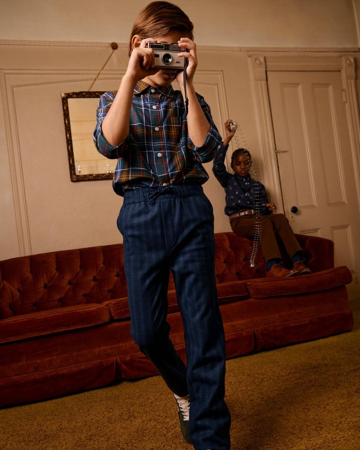 A child wearing Navy Stripe Everyday Stretch Pants poses with a camera, showcasing stylish comfort in a cozy room.