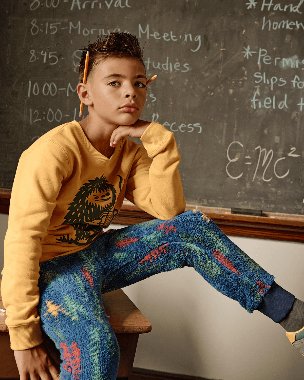 Boy in yellow sweatshirt and Wilderness Highland Fleece Sweatpants sitting on a desk near a blackboard.