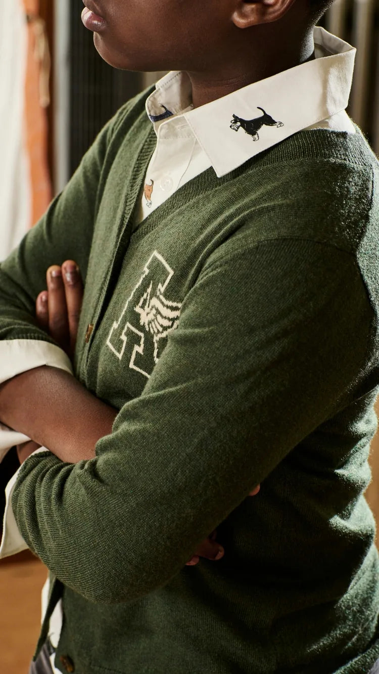 A close-up of a person wearing an olive green knit cardigan over a white collared shirt. The cardigan has a large white embroidered letter 'H' with a black dog silhouette on the chest and a small black dog logo on the collar.