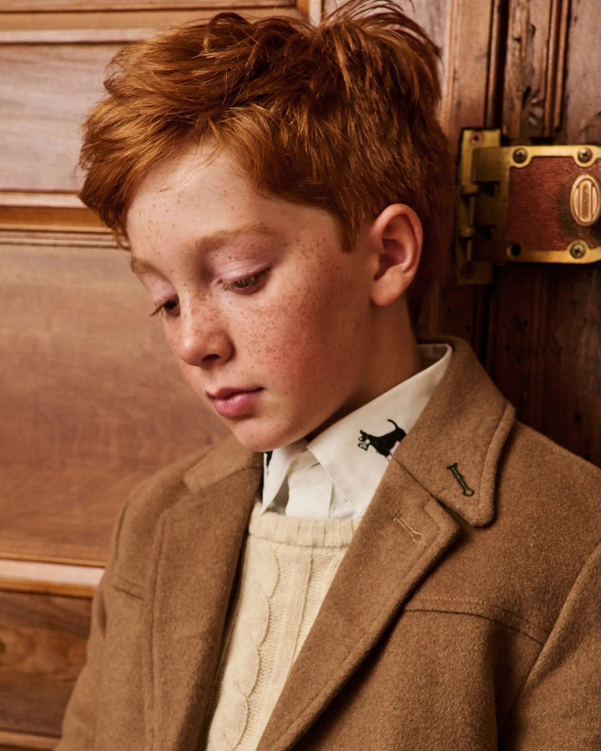 A young boy with red hair and freckles wearing a brown wool coat over a cream sweater and white collared shirt, leaning against a wooden door.