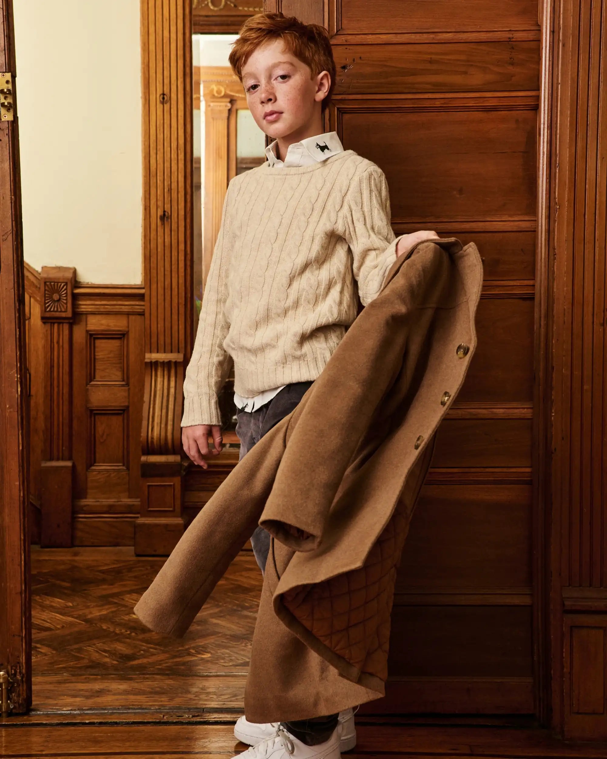 A young boy stands in a warm, wood-paneled room holding a long brown coat. He is wearing a cream cable-knit sweater over a white collared shirt, gray jeans, and white sneakers.