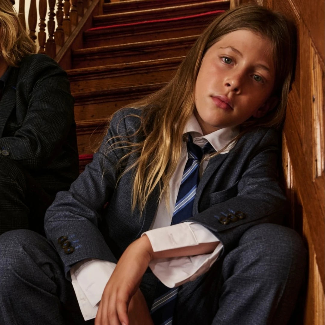 A child model wearing a dark blue textured blazer, matching trousers, a white dress shirt, and a blue striped tie, sitting on a wooden staircase.
