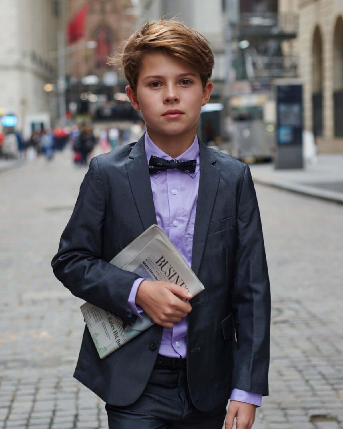A young boy in a stylish suit holding a newspaper, wearing a Lavender Purple Standard Button Down and bow tie.