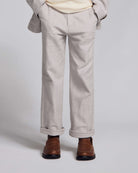 A person shown from the waist down wearing light beige trousers with rolled cuffs, a cream shirt, and brown leather loafers against a light gray studio background.