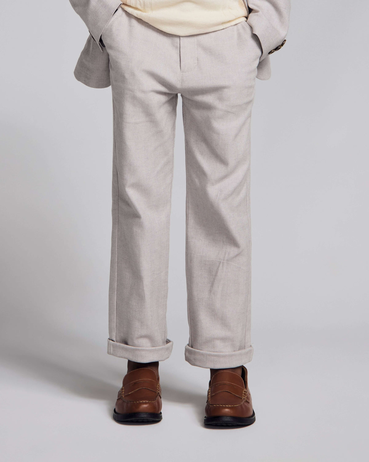 A person shown from the waist down wearing light beige trousers with rolled cuffs, a cream shirt, and brown leather loafers against a light gray studio background.