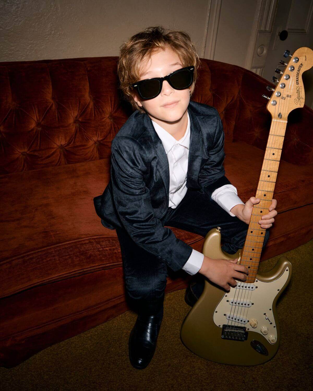Kid wearing a Midnight Velvet Plaid Blazer and sunglasses, posing with a guitar on a couch.