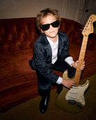 Kid wearing a Midnight Velvet Plaid Blazer and sunglasses, posing with a guitar on a couch.