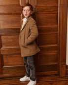 A boy wearing a camel wool coat, standing on a staircase, alternate view. Appaman Fall/Winter 2026 collection.